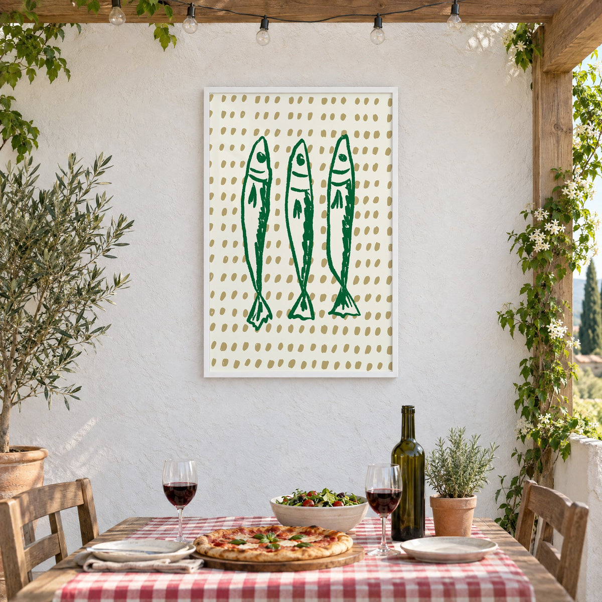 Three green sardines on dotted print in a white frame on a stucco wall above a checked-cloth table with pizza and red wine