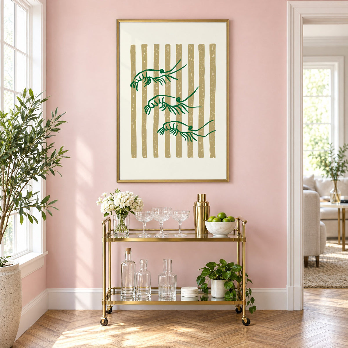Three green shrimp on gold stripes print in a brass frame on a blush pink wall above a gold bar cart with glassware