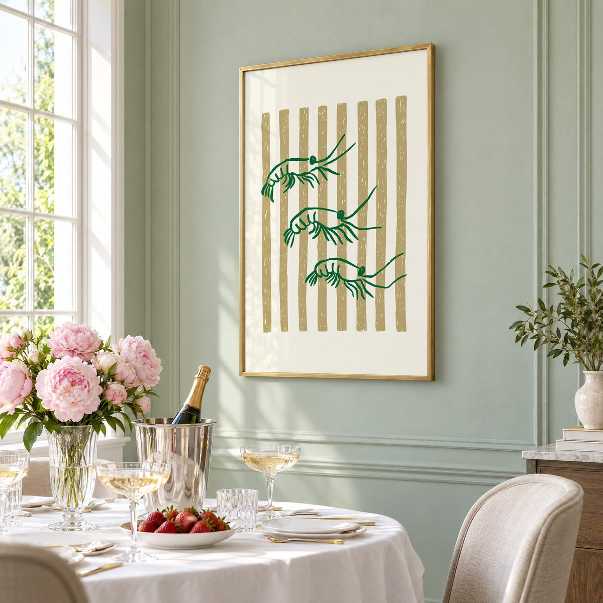 Three green shrimp on gold stripes print in a brass frame on a sage wall above a brunch table with champagne and peonies, seen at an angle