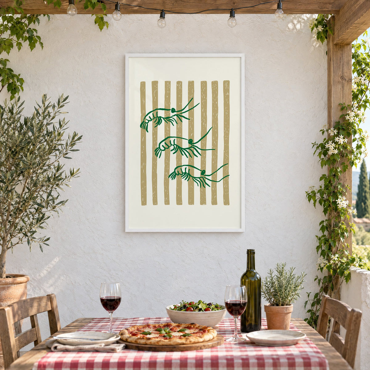 Three green shrimp on gold stripes print in a white frame on a stucco wall above a checked-cloth table with pizza and red wine