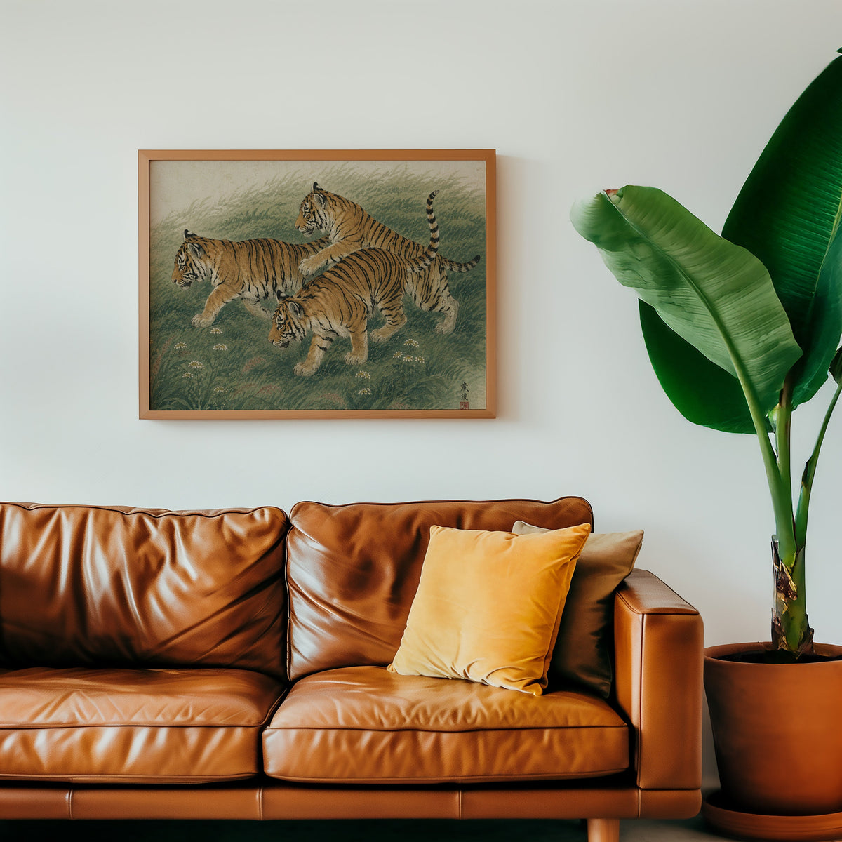 Three tiger cubs in tall grass print in a warm oak frame above a cognac leather sofa with a mustard pillow
