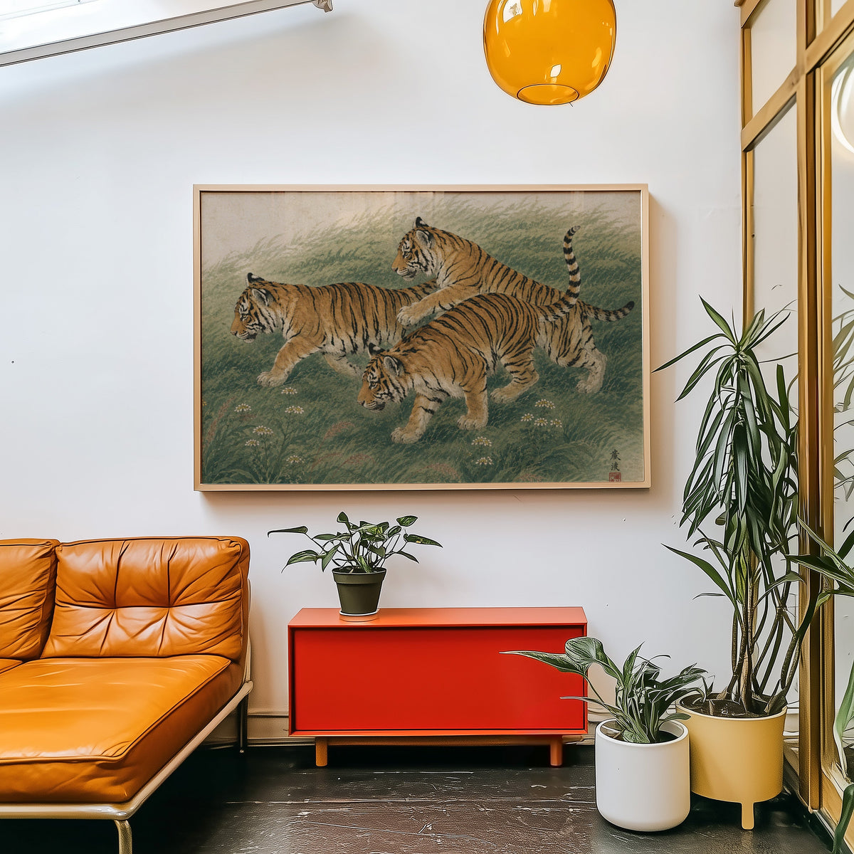 Three tiger cubs in tall grass print in a light wood frame above a red cabinet beside a tan leather daybed