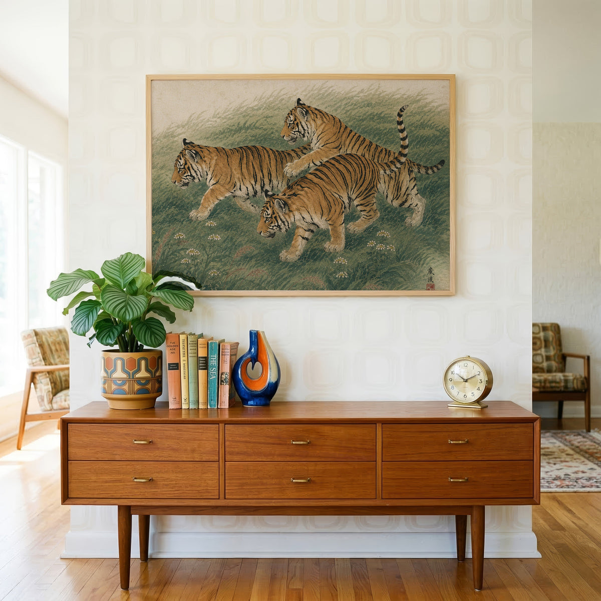 Three tiger cubs in tall grass print in a light oak frame above a teak credenza with books and a brass clock