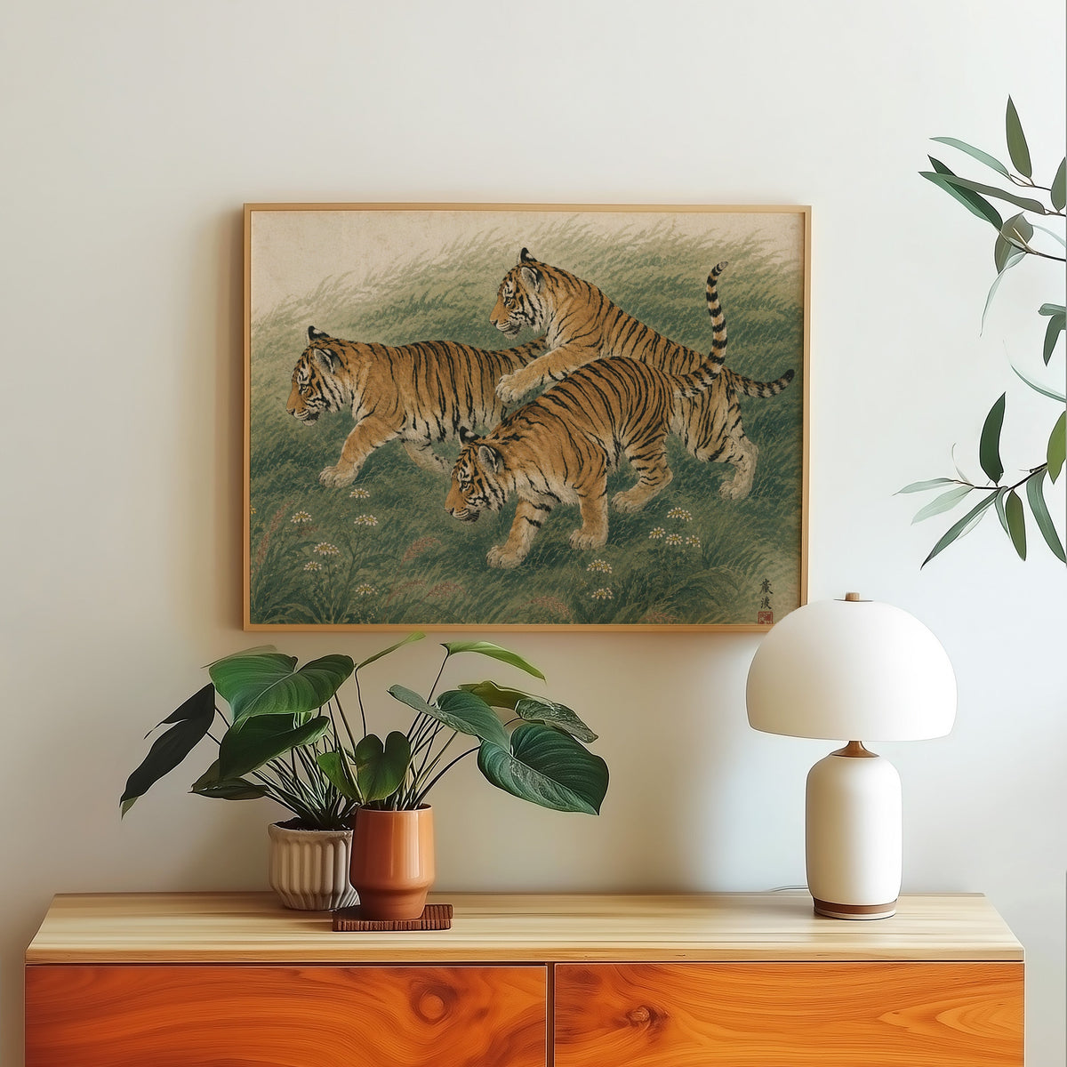 Three tiger cubs in tall grass print in a light oak frame above a wood sideboard with a white dome lamp