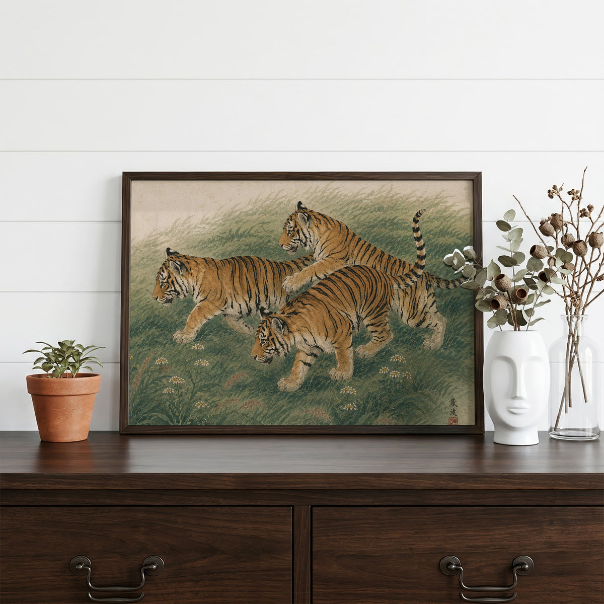 Three tiger cubs in tall grass print in a dark walnut frame on a dark wood dresser beside a white face vase