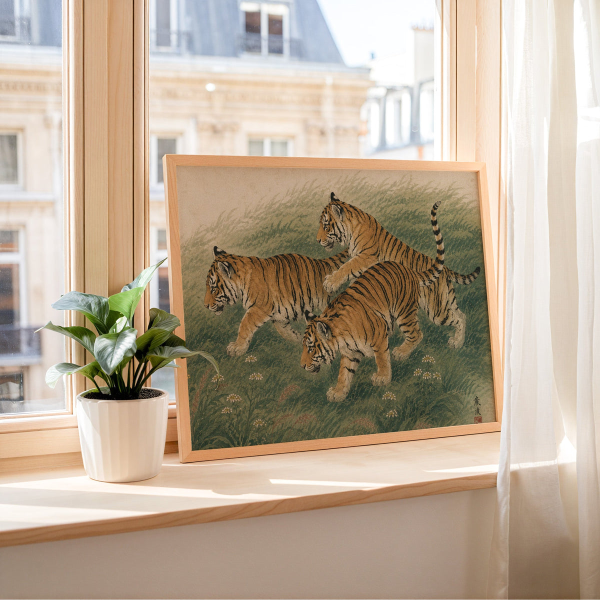 Three tiger cubs in tall grass print in a light wood frame leaning on a sunny windowsill by sheer curtains