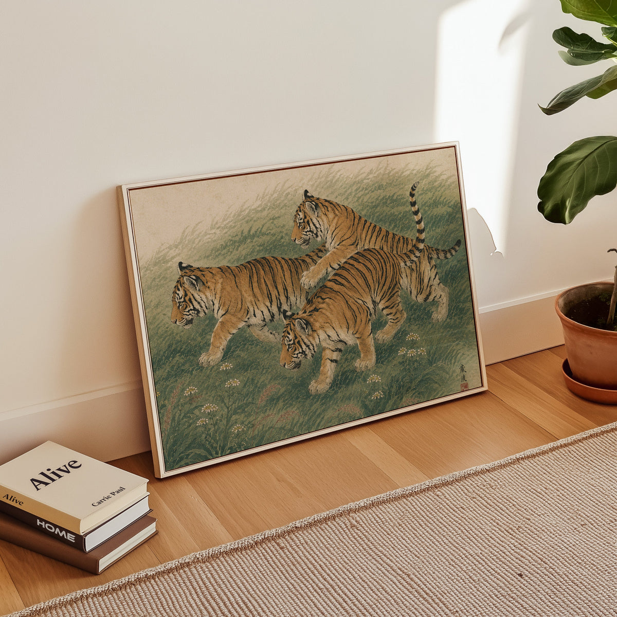 Three tiger cubs in tall grass print in a white floater frame leaning on a wood floor beside stacked books