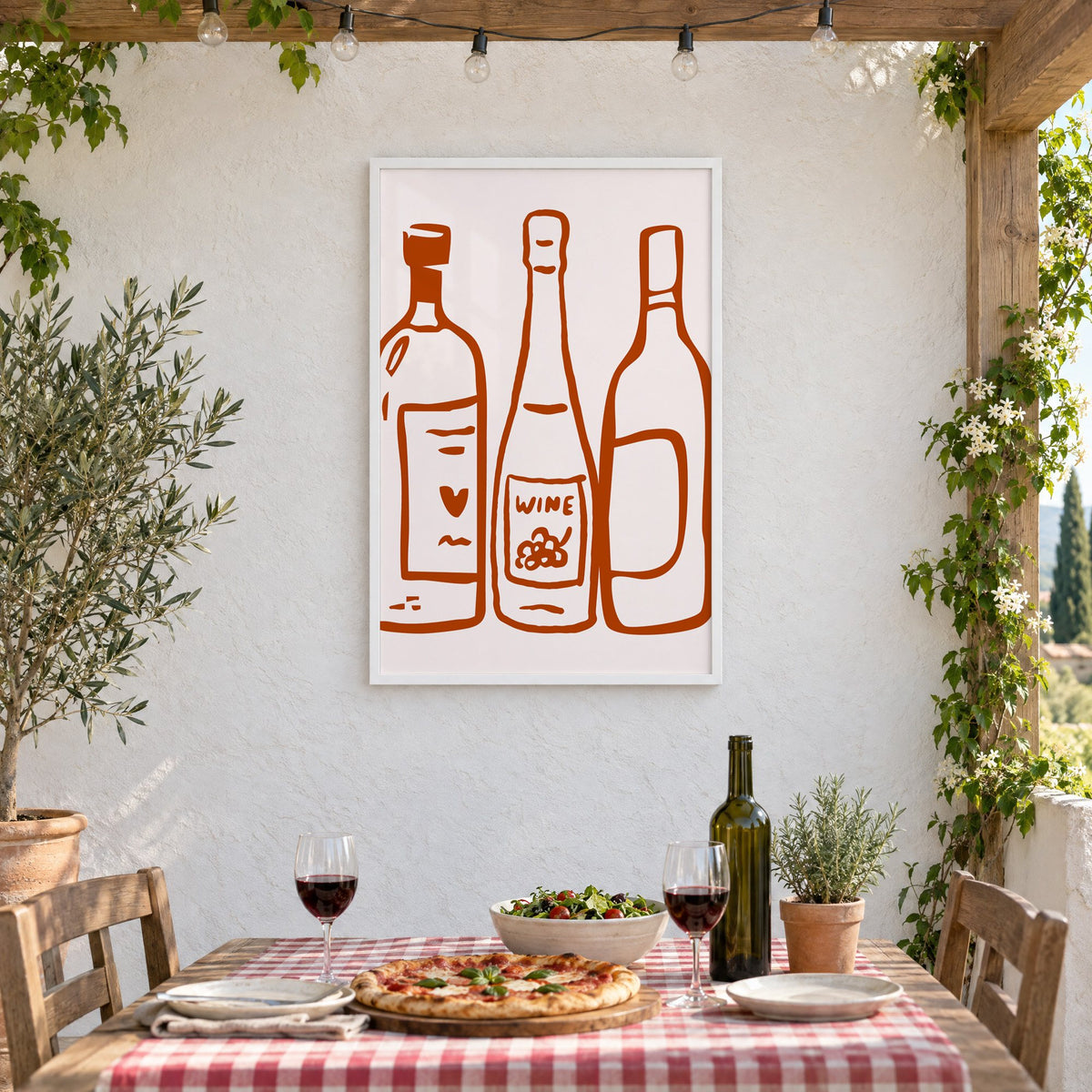 Three wine bottles rust brush line print in a white frame on a stucco wall above a checked-cloth table with pizza and red wine