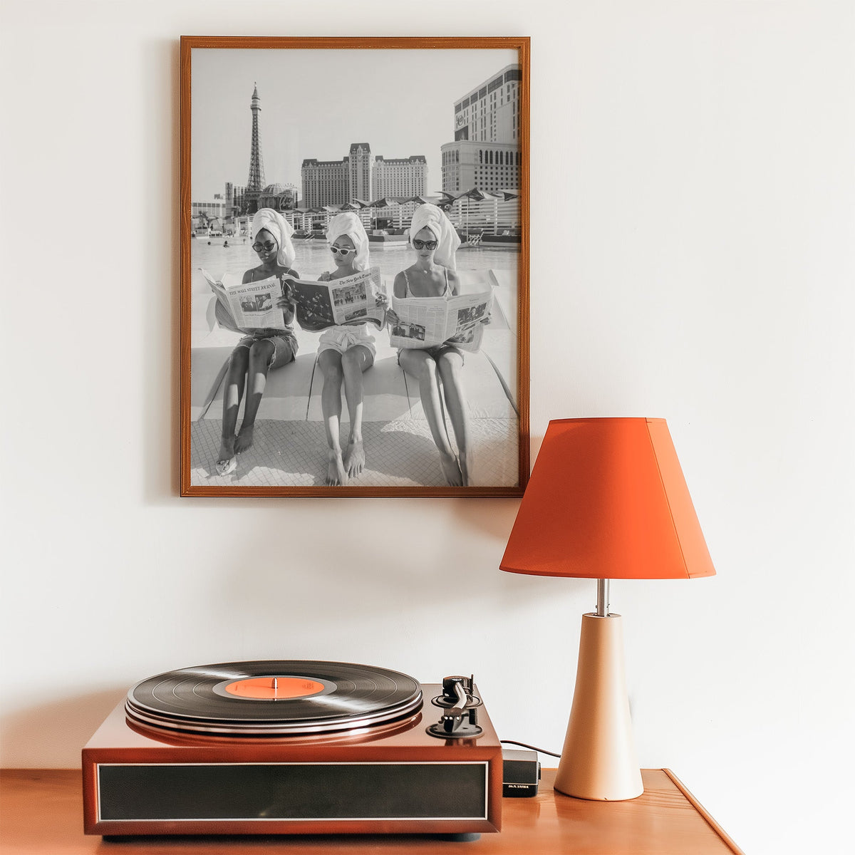 Three women in towel turbans reading newspapers print in a walnut frame above a record player and orange lamp