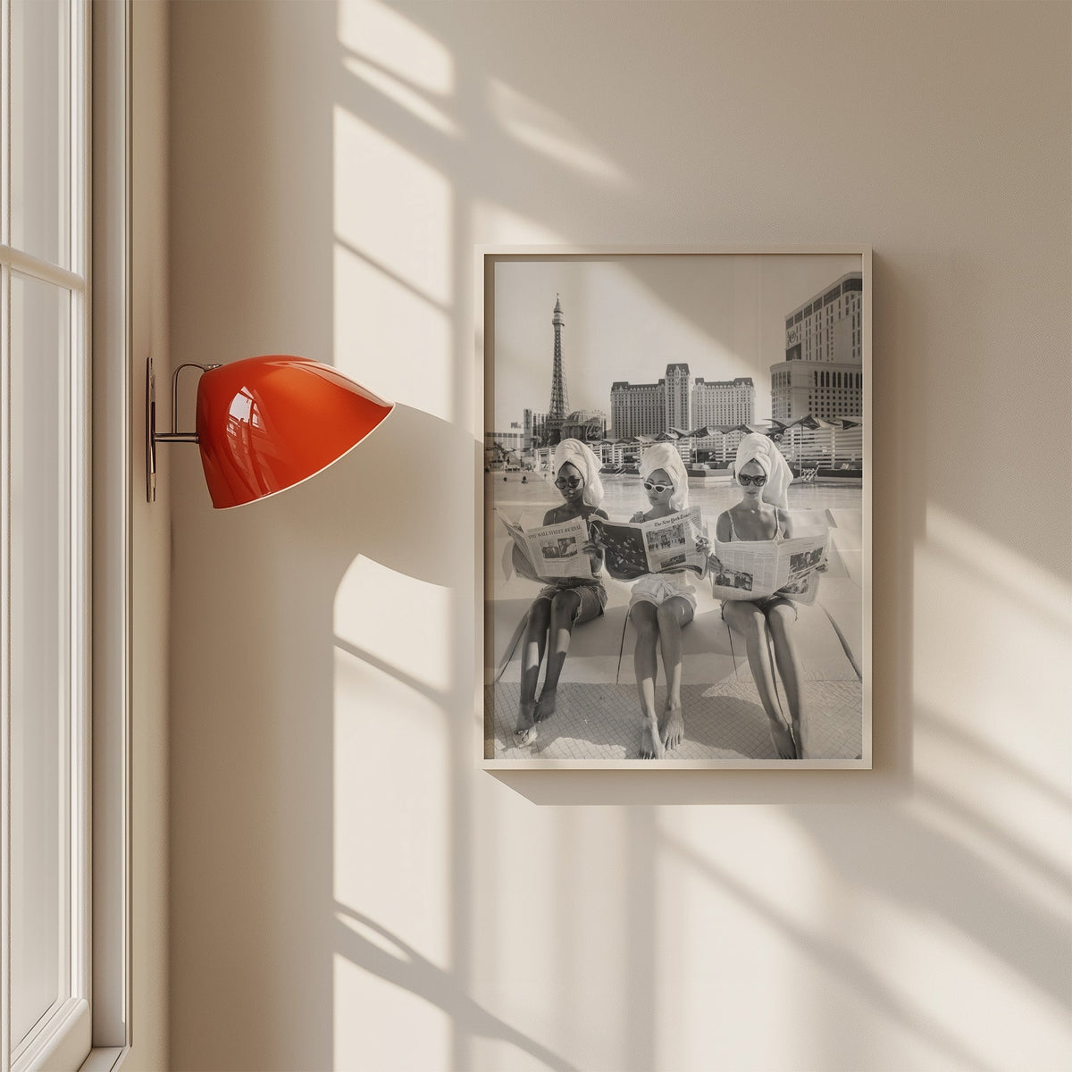 Three women in towel turbans reading newspapers print in a white frame on a sunlit wall beside a red sconce