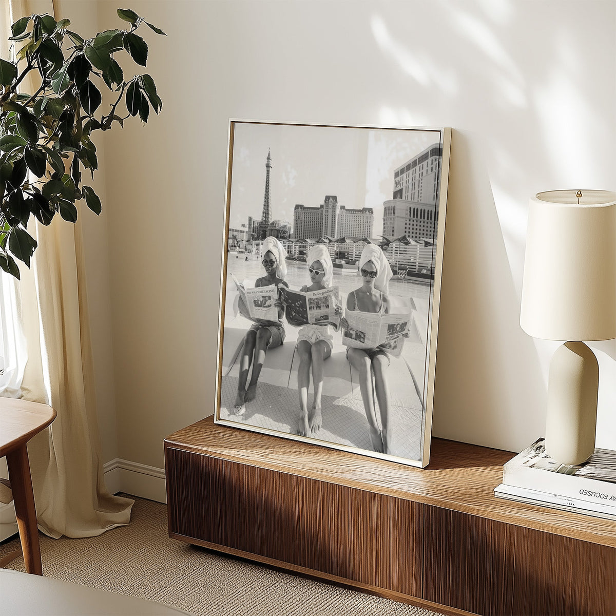 Three women in towel turbans reading newspapers print in a white frame on a walnut sideboard beside a ceramic lamp