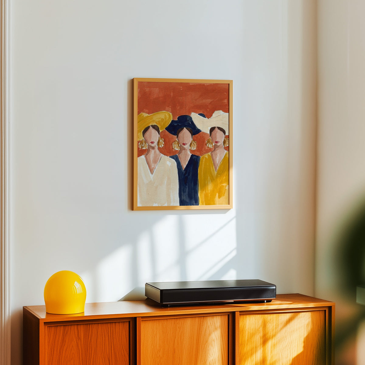 Three friends fashion print in a light oak frame above a teak sideboard with a yellow lamp