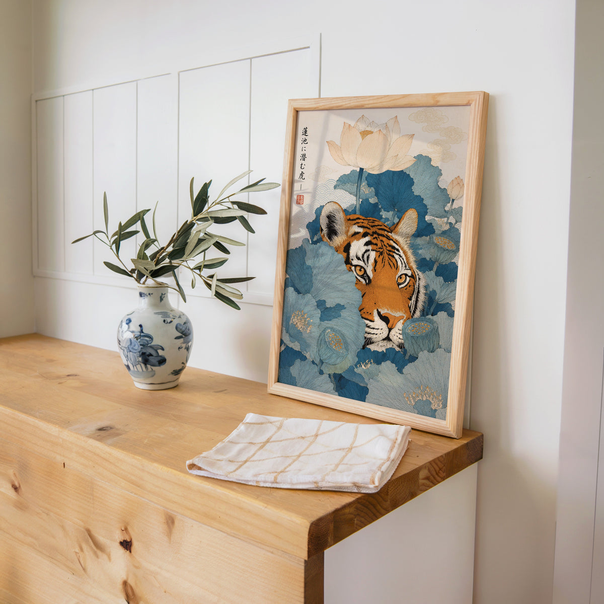 tiger hiding in blue lotus leaves print in a pine frame on a wooden counter beside a blue and white porcelain vase