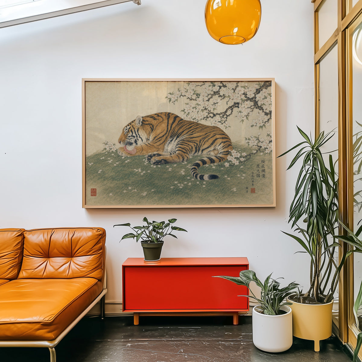 Tiger resting under cherry blossoms print in a light wood frame above a red cabinet beside a tan leather daybed