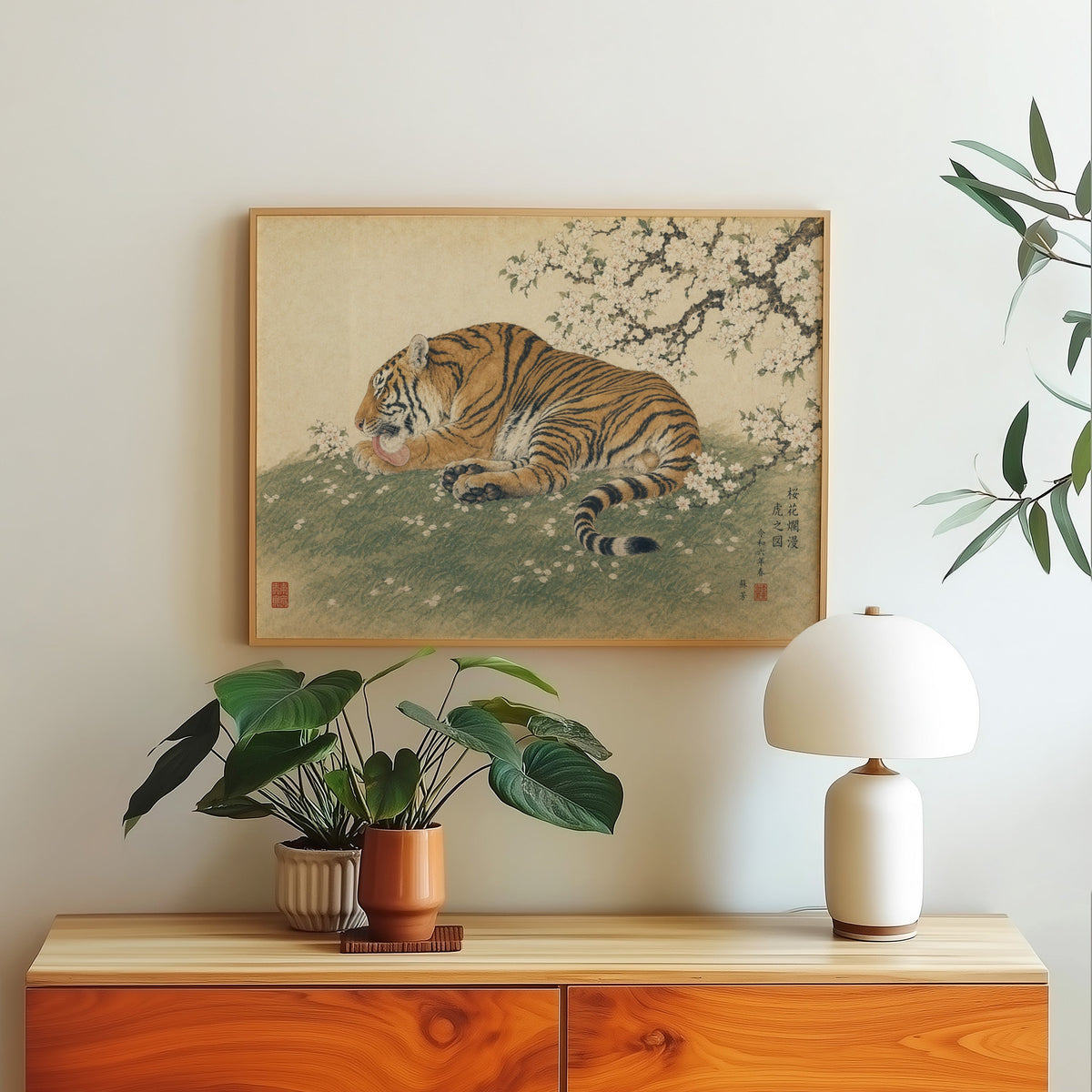 Tiger resting under cherry blossoms print in a light oak frame above a wood sideboard with a white dome lamp
