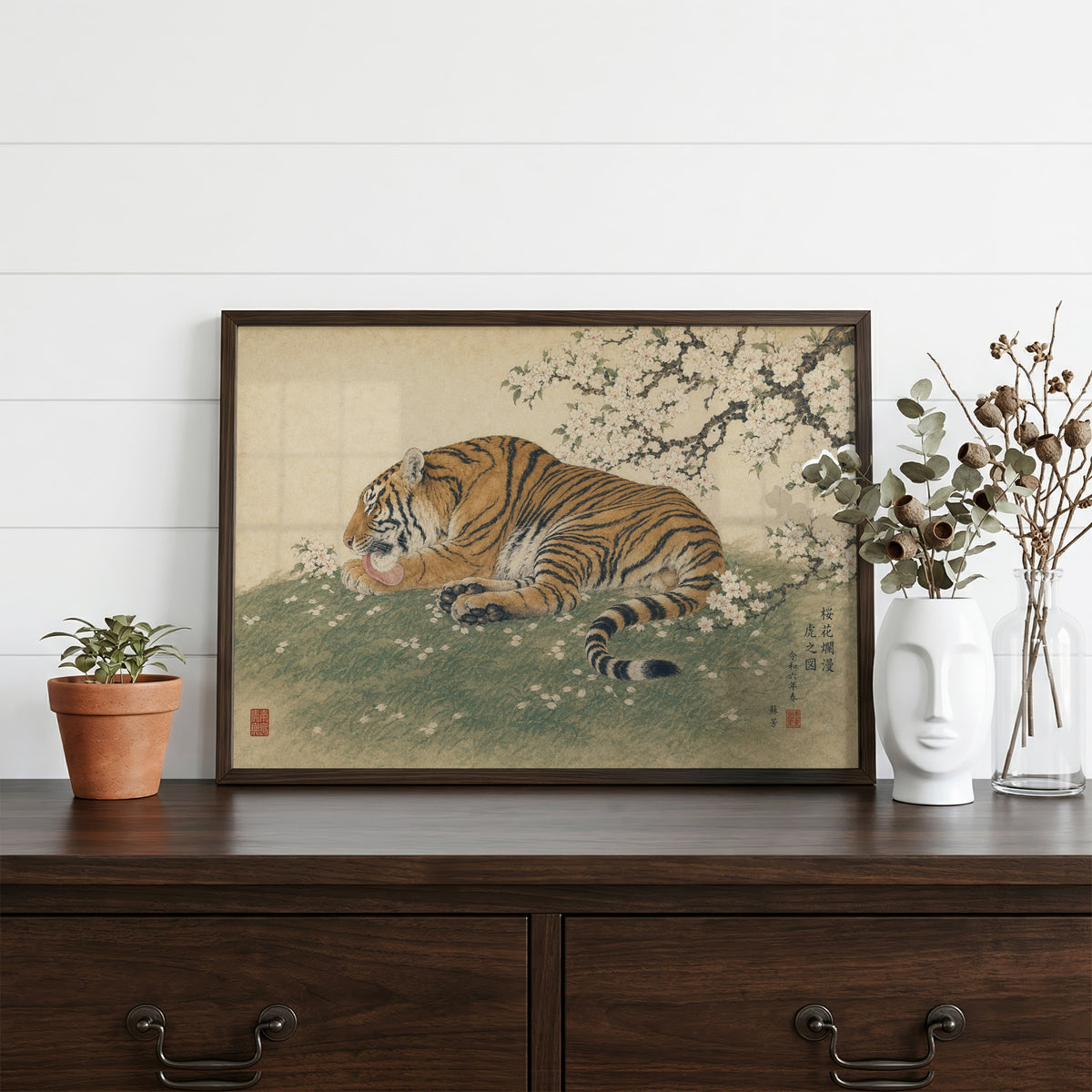 Tiger resting under cherry blossoms print in a dark walnut frame on a dark wood dresser beside a white face vase