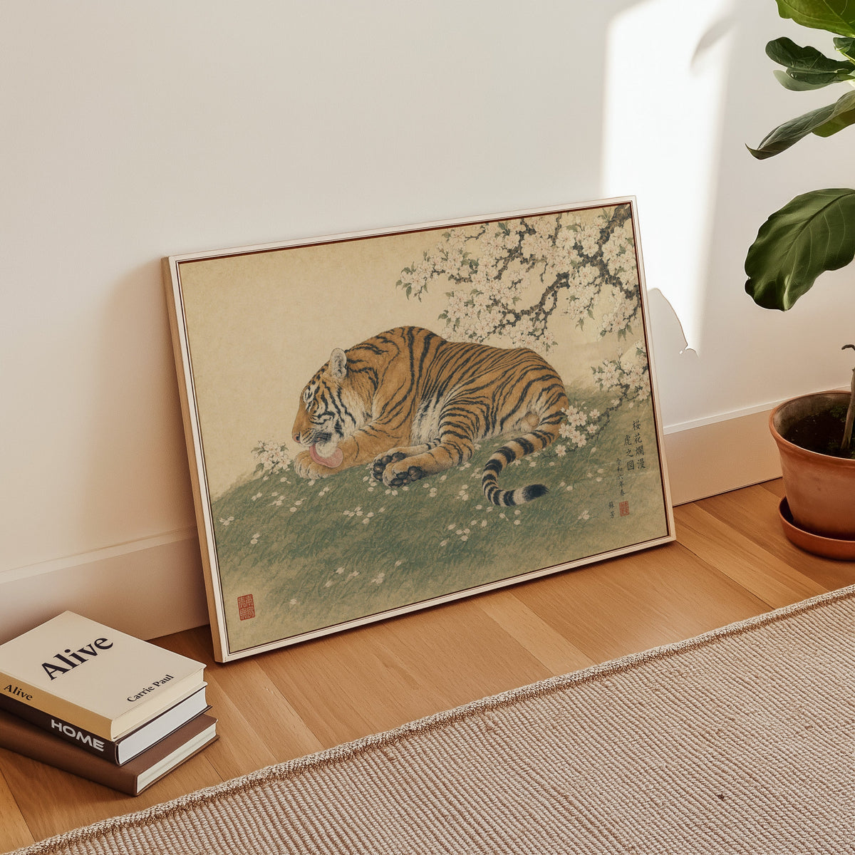Tiger resting under cherry blossoms print in a white floater frame leaning on a wood floor beside stacked books