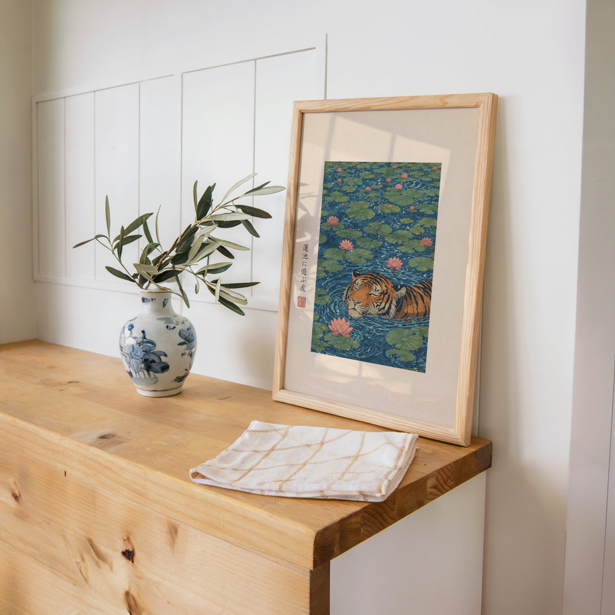 tiger swimming in lotus pond print in a pine frame on a wooden counter beside a blue and white porcelain vase