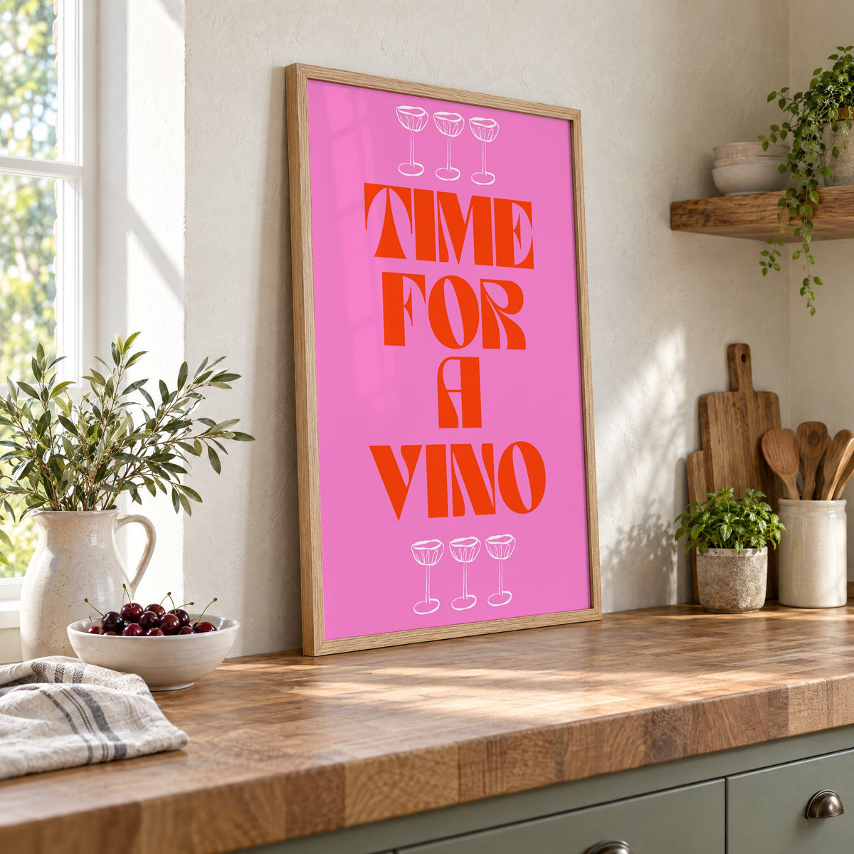 Time for a Vino pink coupes print in an oak frame leaning on a butcher block island beside a bowl of cherries