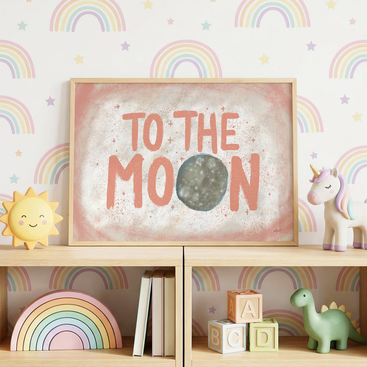 To the moon coral lettering nursery print in an oak frame on a toy shelf against rainbow wallpaper