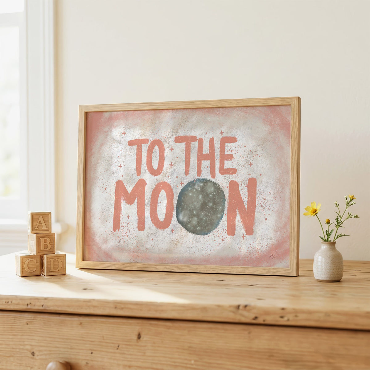 To the moon coral lettering nursery print in an oak frame on a wooden dresser beside ABC blocks