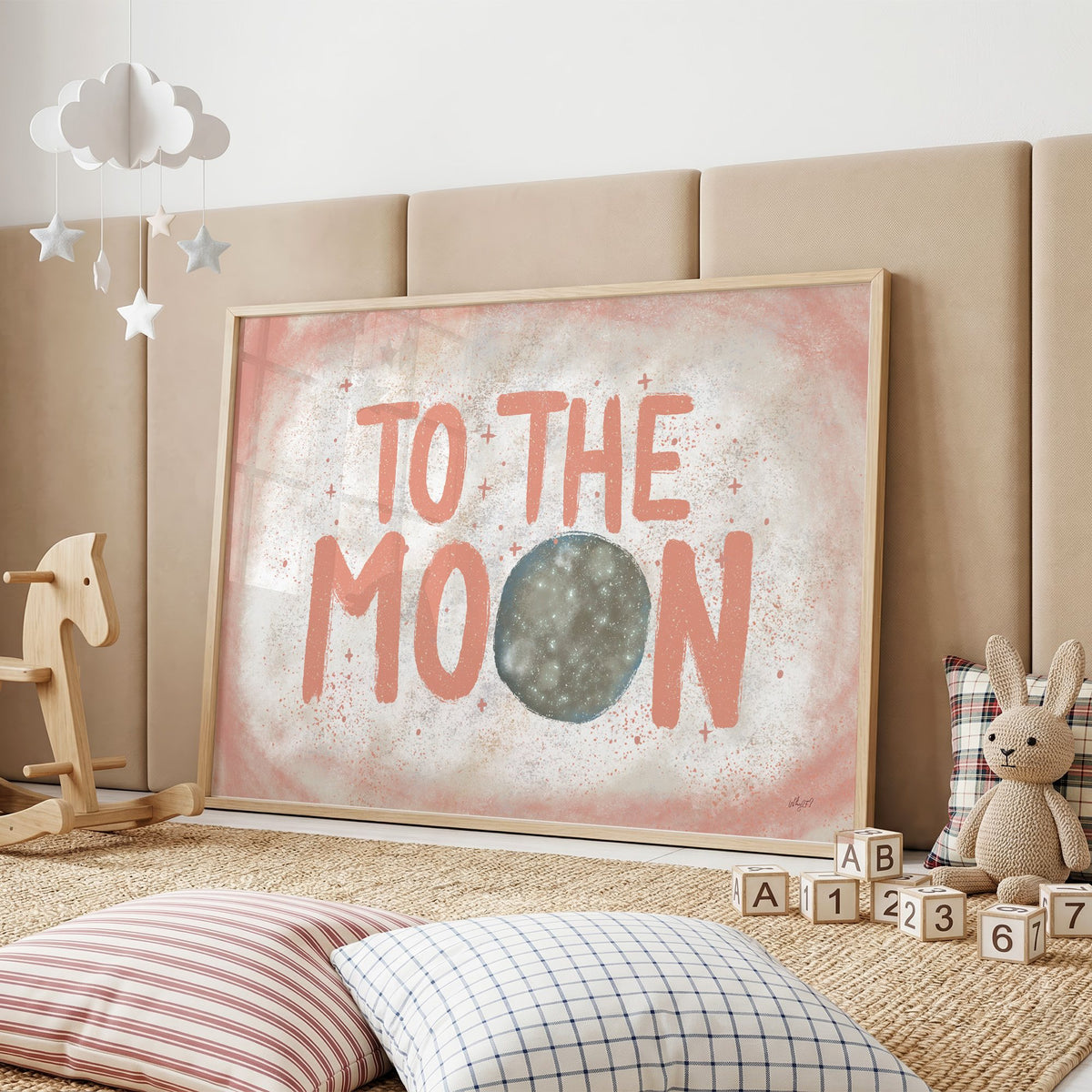 To the moon coral lettering nursery print in an oak frame on a padded wall by a wooden rocking horse