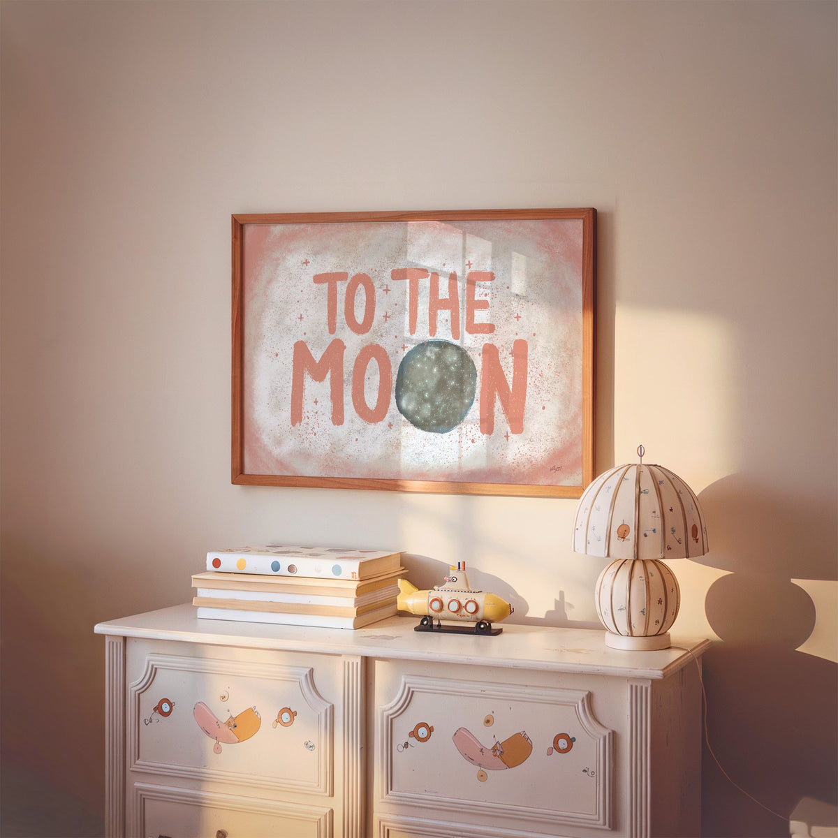 To the moon coral lettering nursery print in a cherry wood frame above a painted dresser and lamp