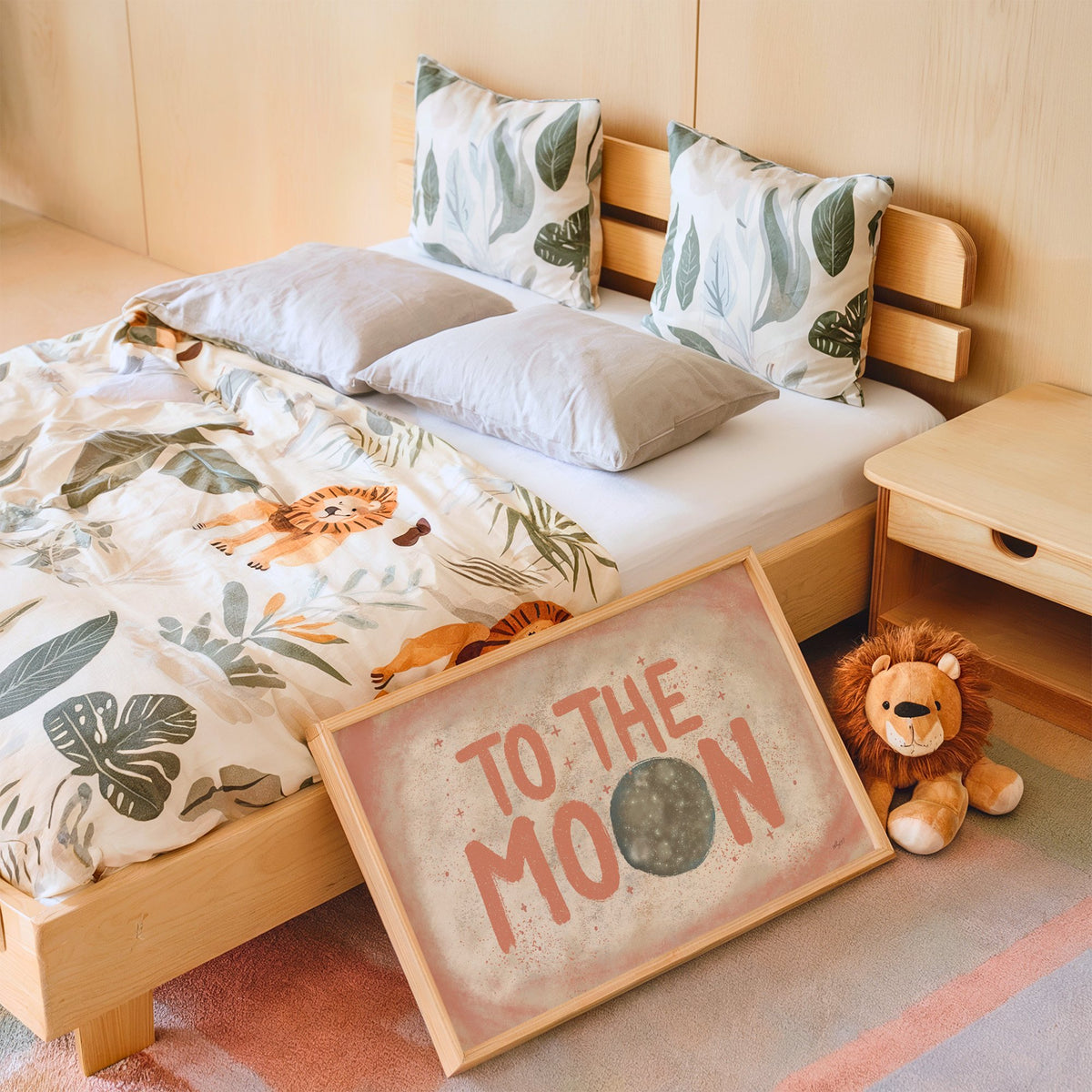 To the moon coral lettering nursery print in an oak frame leaning on a kids bed by a lion plush