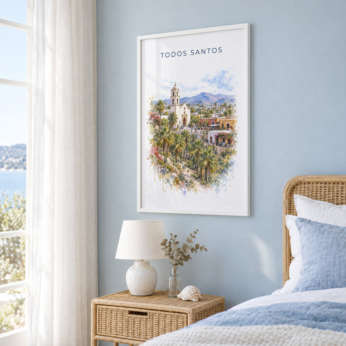 Todos Santos watercolor print in a white frame on a pale blue wall beside a rattan bed in a coastal bedroom