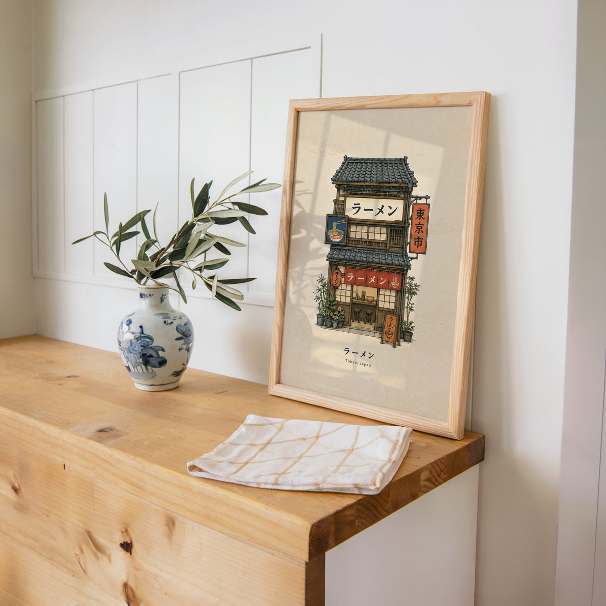 Tokyo ramen shop storefront print in a pine frame on a wooden counter beside a blue and white porcelain vase