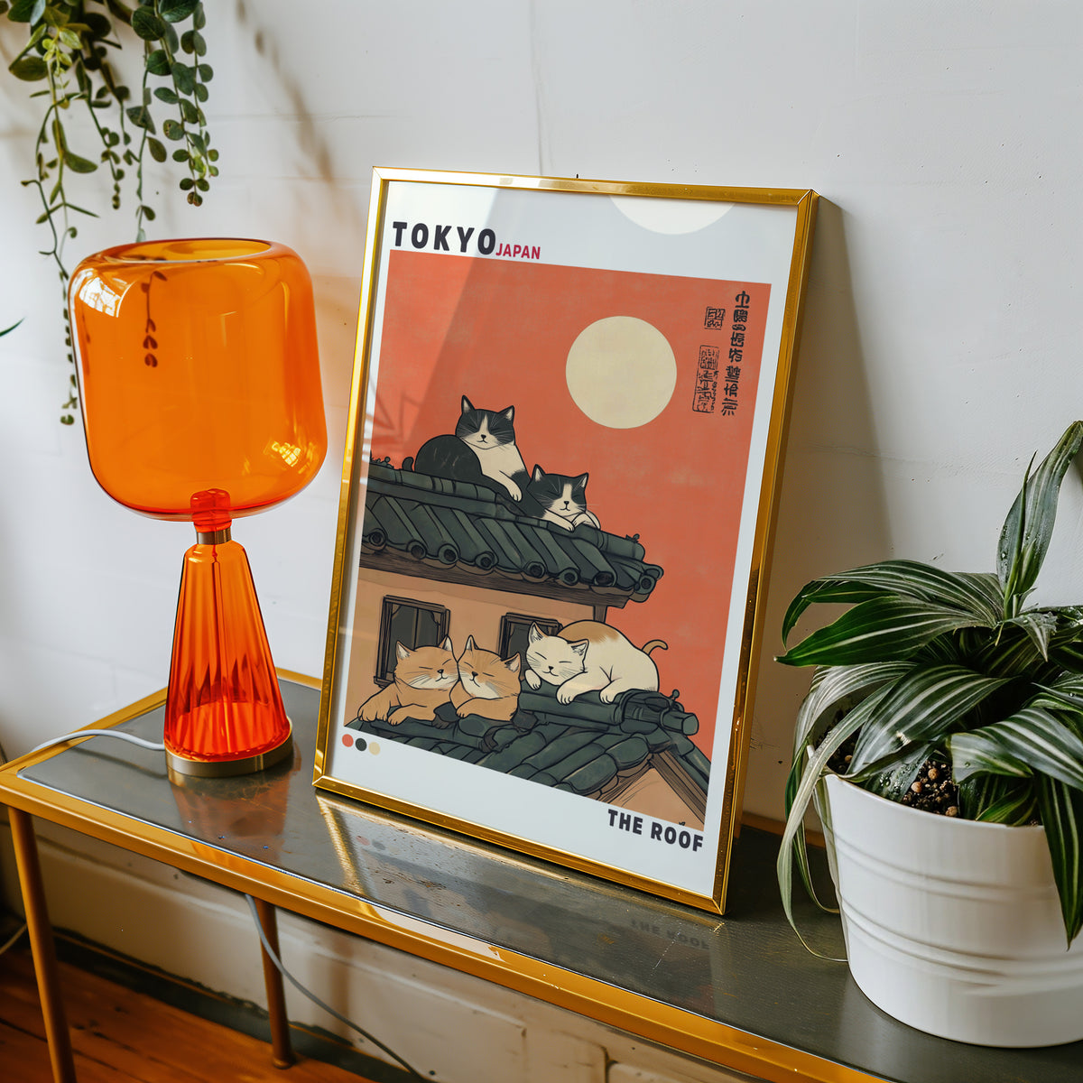 Five cats sleeping on Tokyo roof print shown in a gold frame on a console beside an orange glass lamp