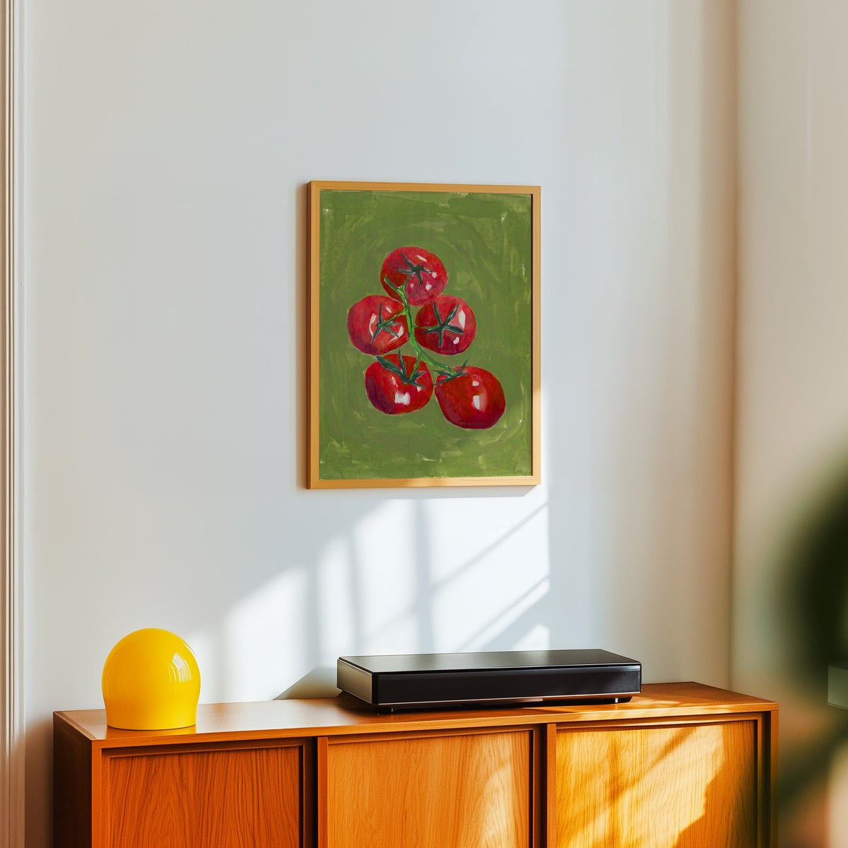 Tomato still life print in a light oak frame above a teak sideboard with a yellow lamp