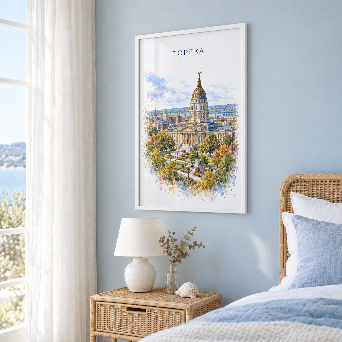 Topeka watercolor print in a white frame on a pale blue wall beside a rattan bed in a coastal bedroom