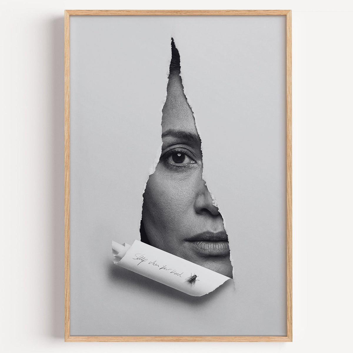Half face behind torn grey paper slit print in a light oak frame on a plain white wall