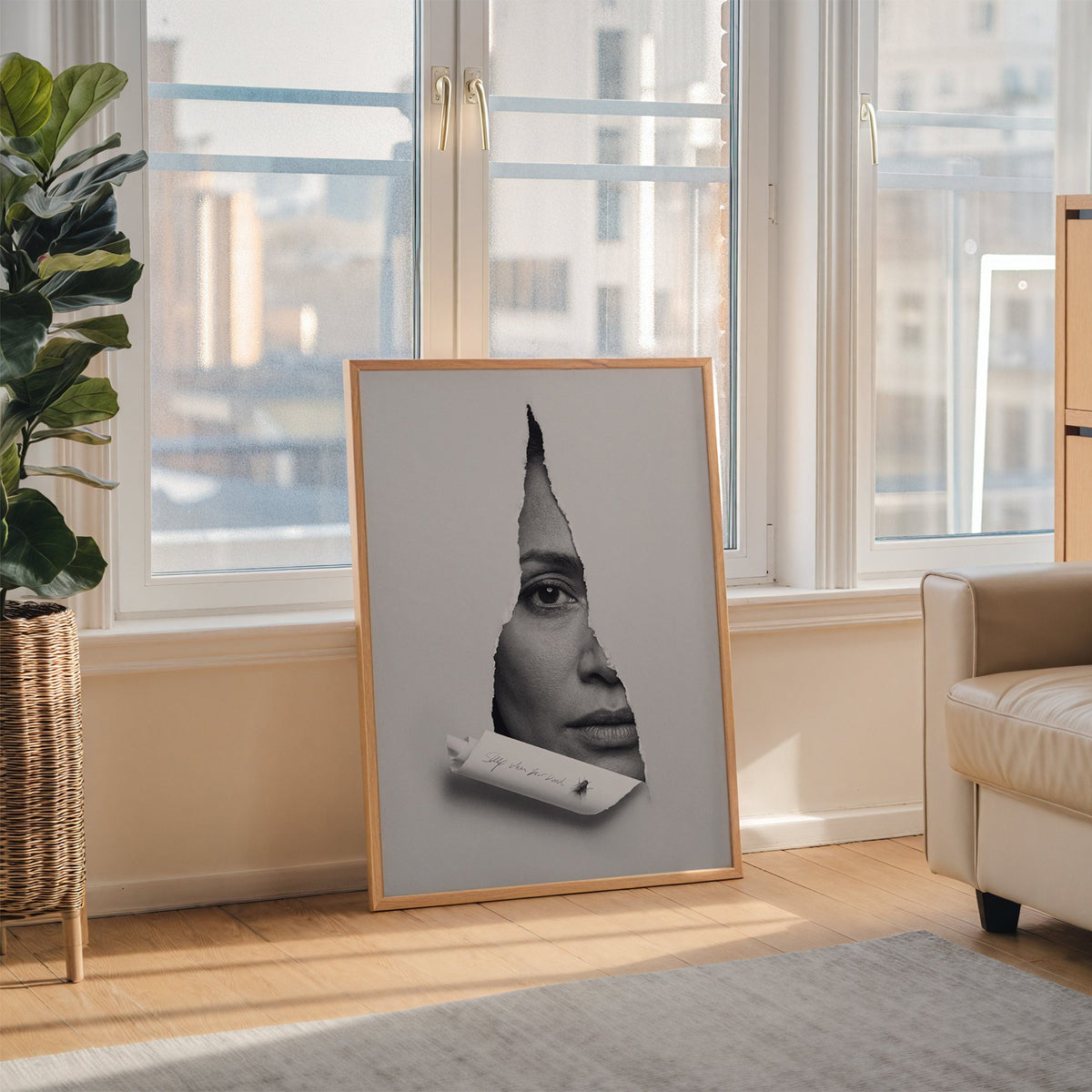 Half face behind torn grey paper slit print in an oak frame leaning on the floor beside a fiddle leaf fig