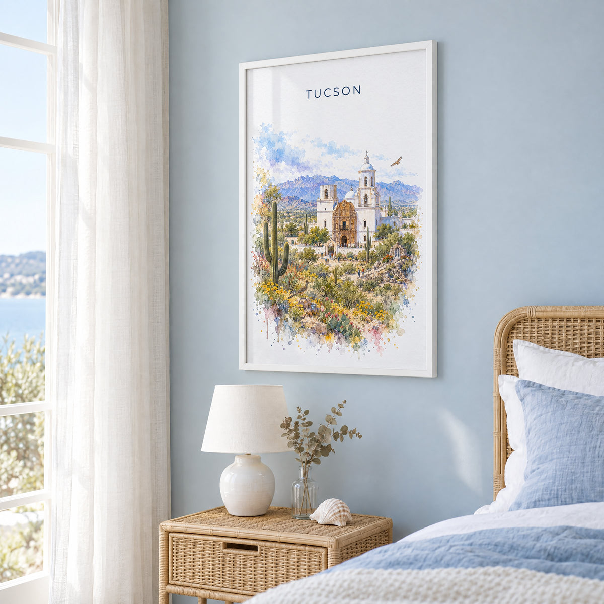 Tucson watercolor print in a white frame on a pale blue wall beside a rattan bed in a coastal bedroom