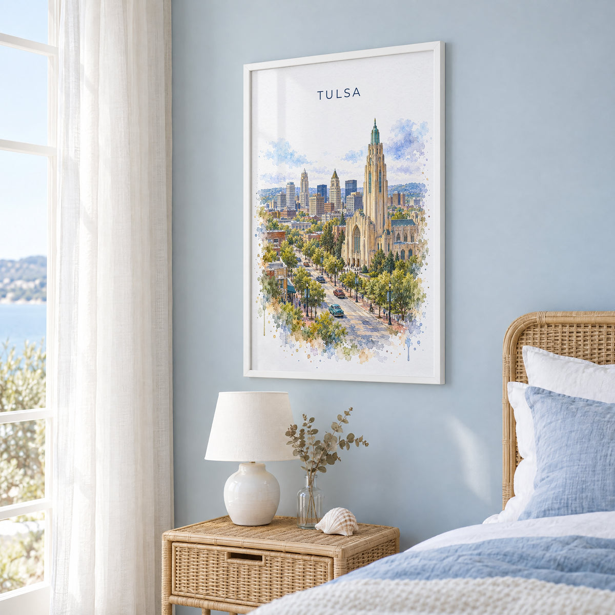 Tulsa watercolor print in a white frame on a pale blue wall beside a rattan bed in a coastal bedroom