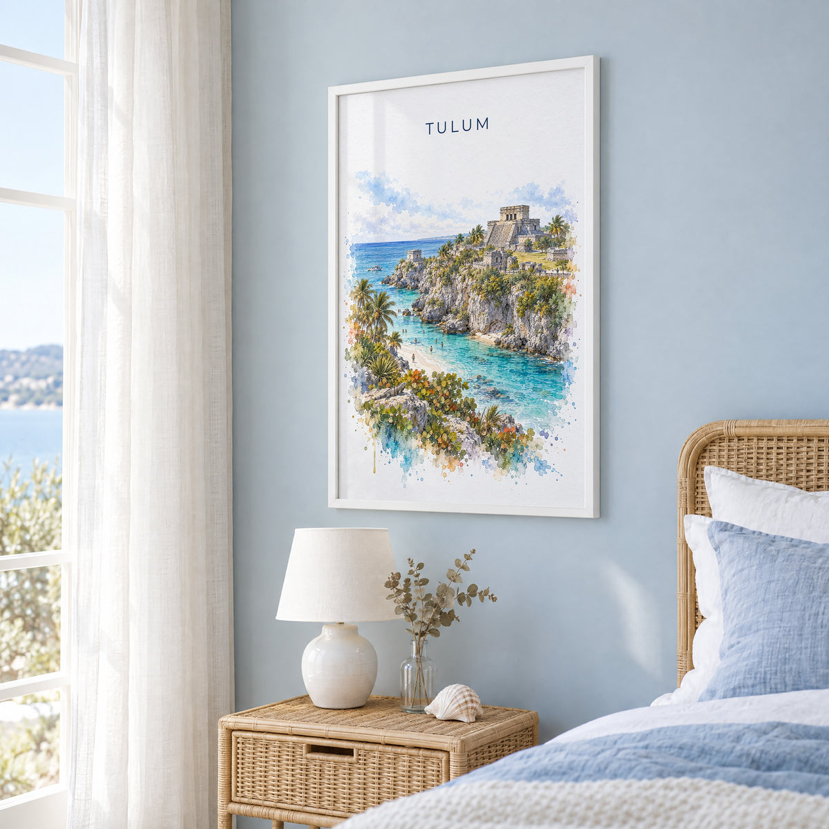 Tulum watercolor print in a white frame on a pale blue wall beside a rattan bed in a coastal bedroom