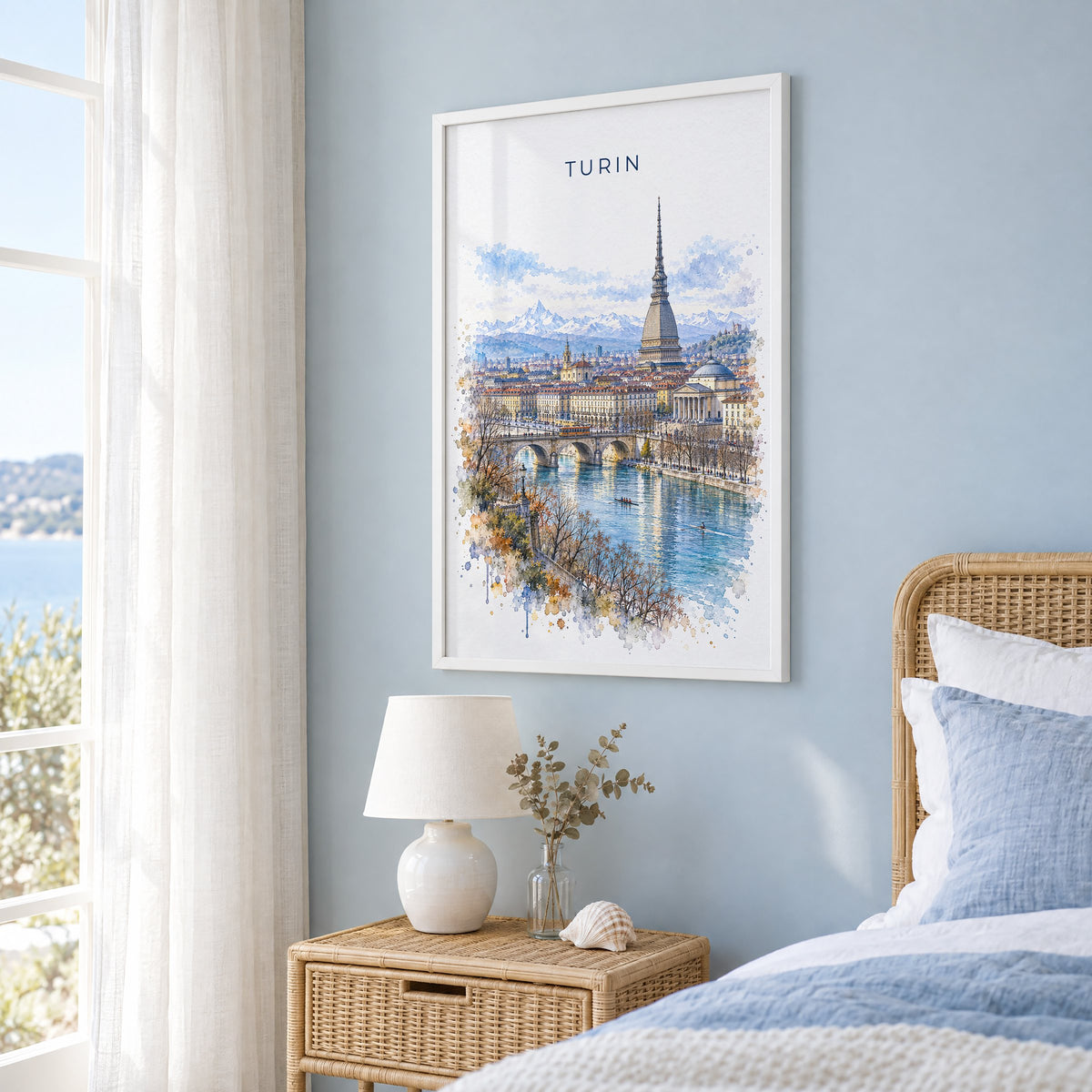 Turin watercolor print in a white frame on a pale blue wall beside a rattan bed in a coastal bedroom