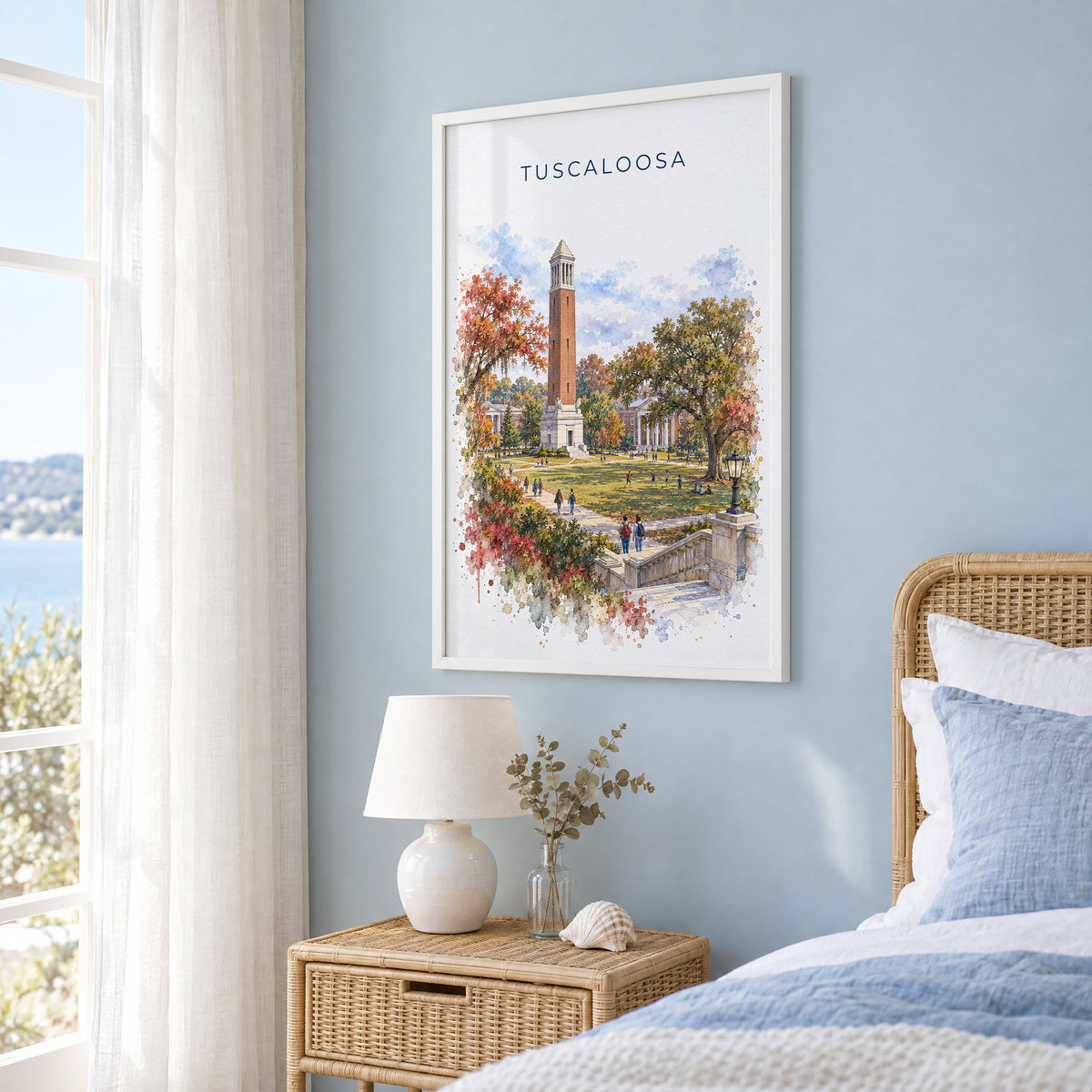 Tuscaloosa watercolor print in a white frame on a pale blue wall beside a rattan bed in a coastal bedroom