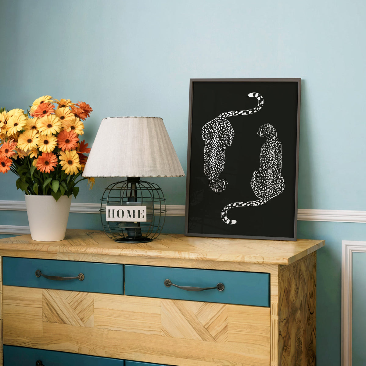 Twin cheetahs in white line art on black print in a charcoal frame on a teal dresser beside orange daisies