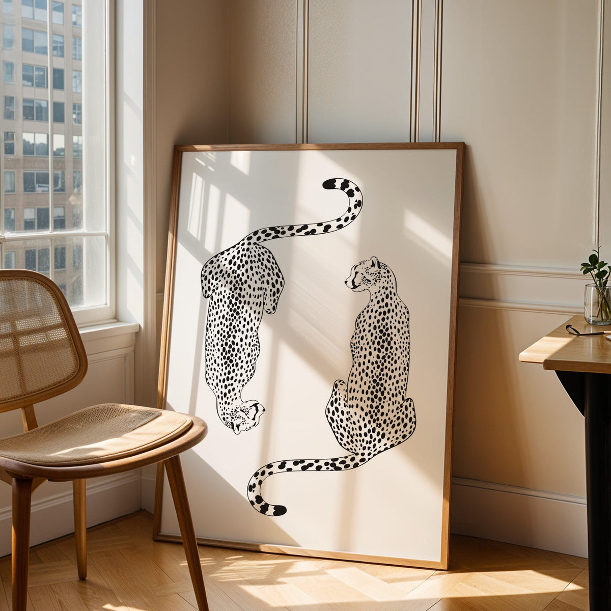 Two black line art cheetahs on white print in an oak frame leaning in a sunlit corner beside a cane chair