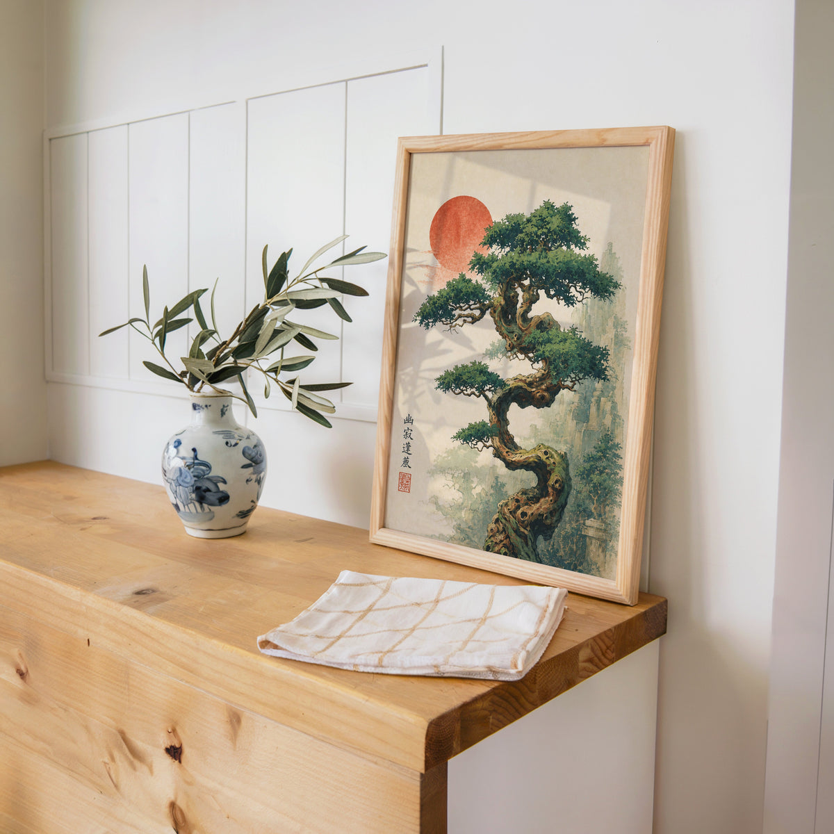 twisted bonsai tree with red sun print in a pine frame on a wooden counter beside a blue and white porcelain vase