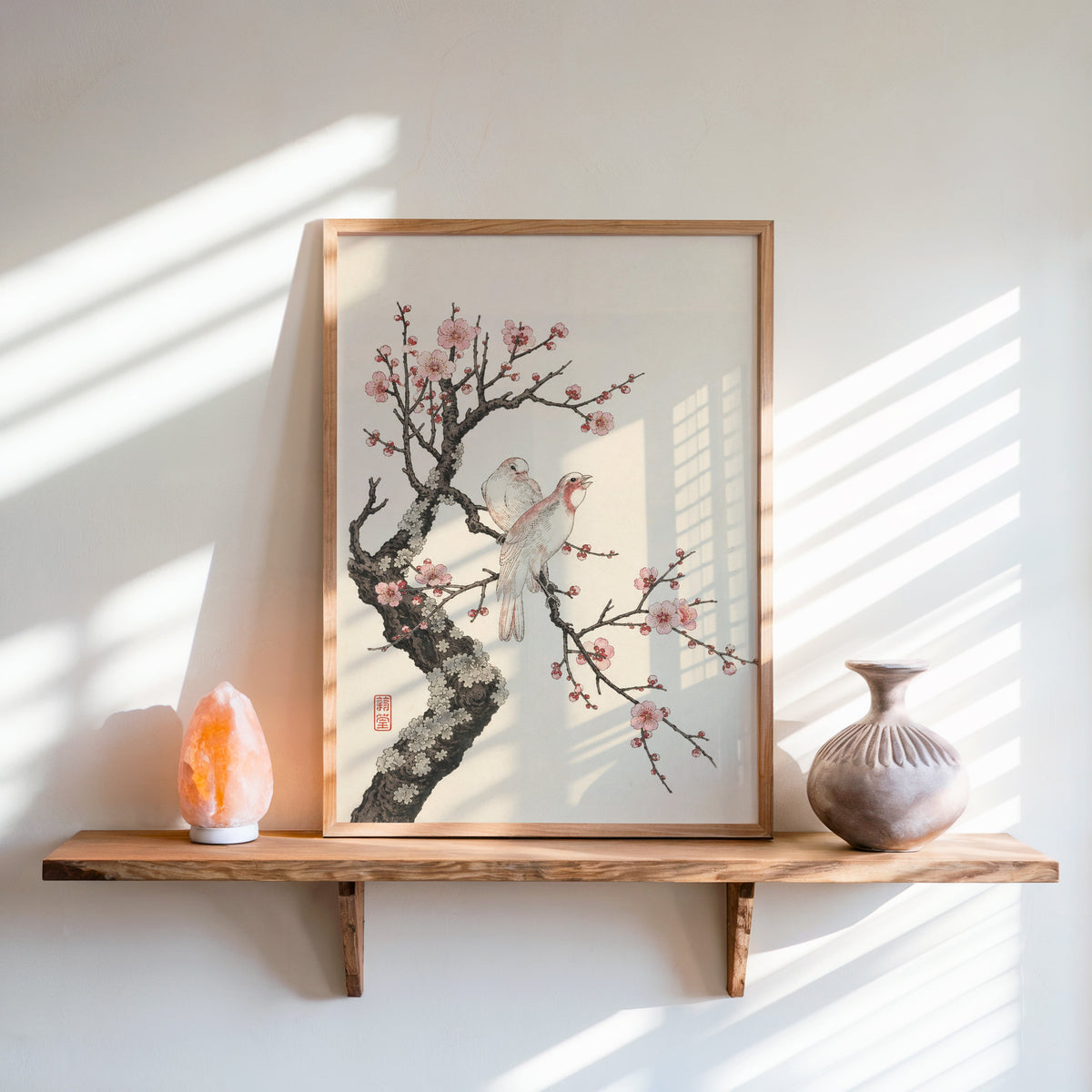two birds on pink blossom branch print in an oak frame on a live-edge wall shelf with a salt lamp and clay vase