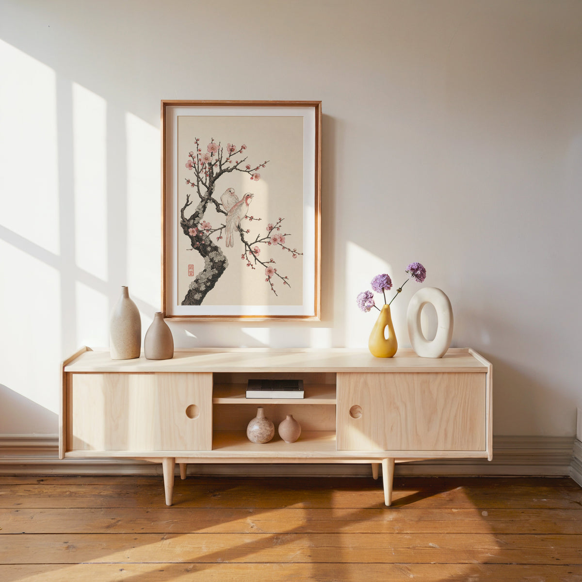 two birds on pink blossom branch print shown in a wood frame with a white mat above a pale wood media console