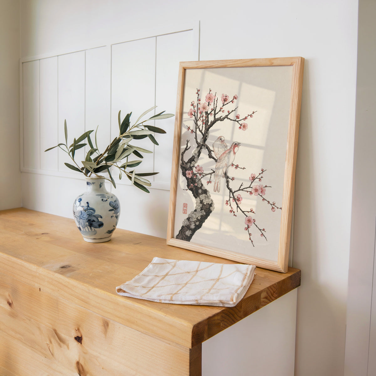 two birds on pink blossom branch print in a pine frame on a wooden counter beside a blue and white porcelain vase