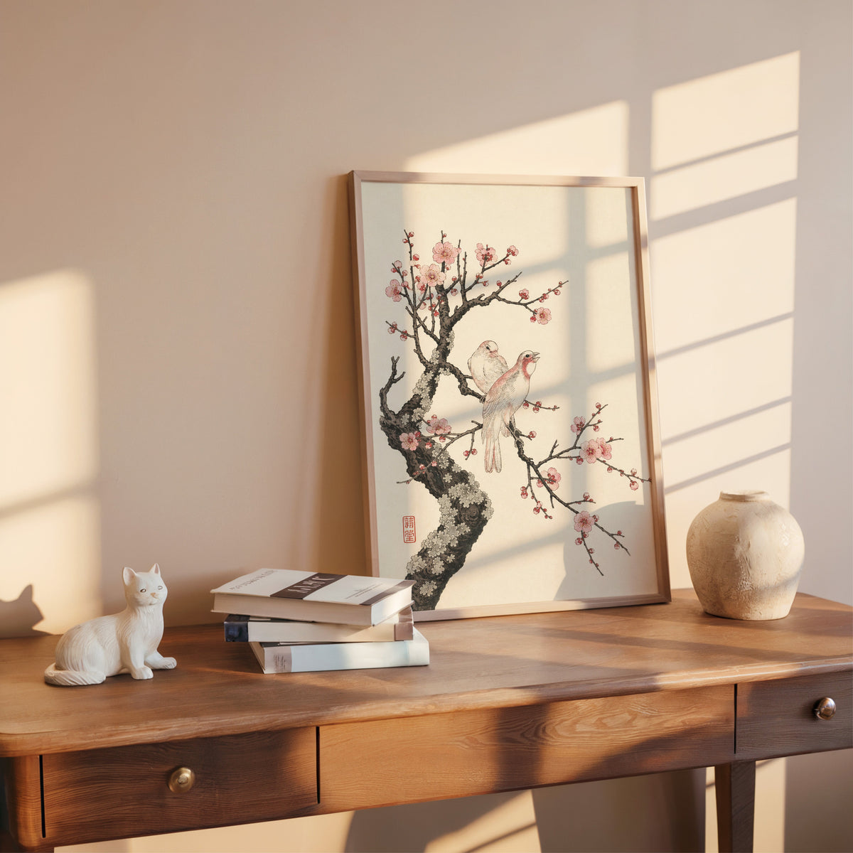 two birds on pink blossom branch print shown in a rose gold frame leaning on a wooden writing desk with books