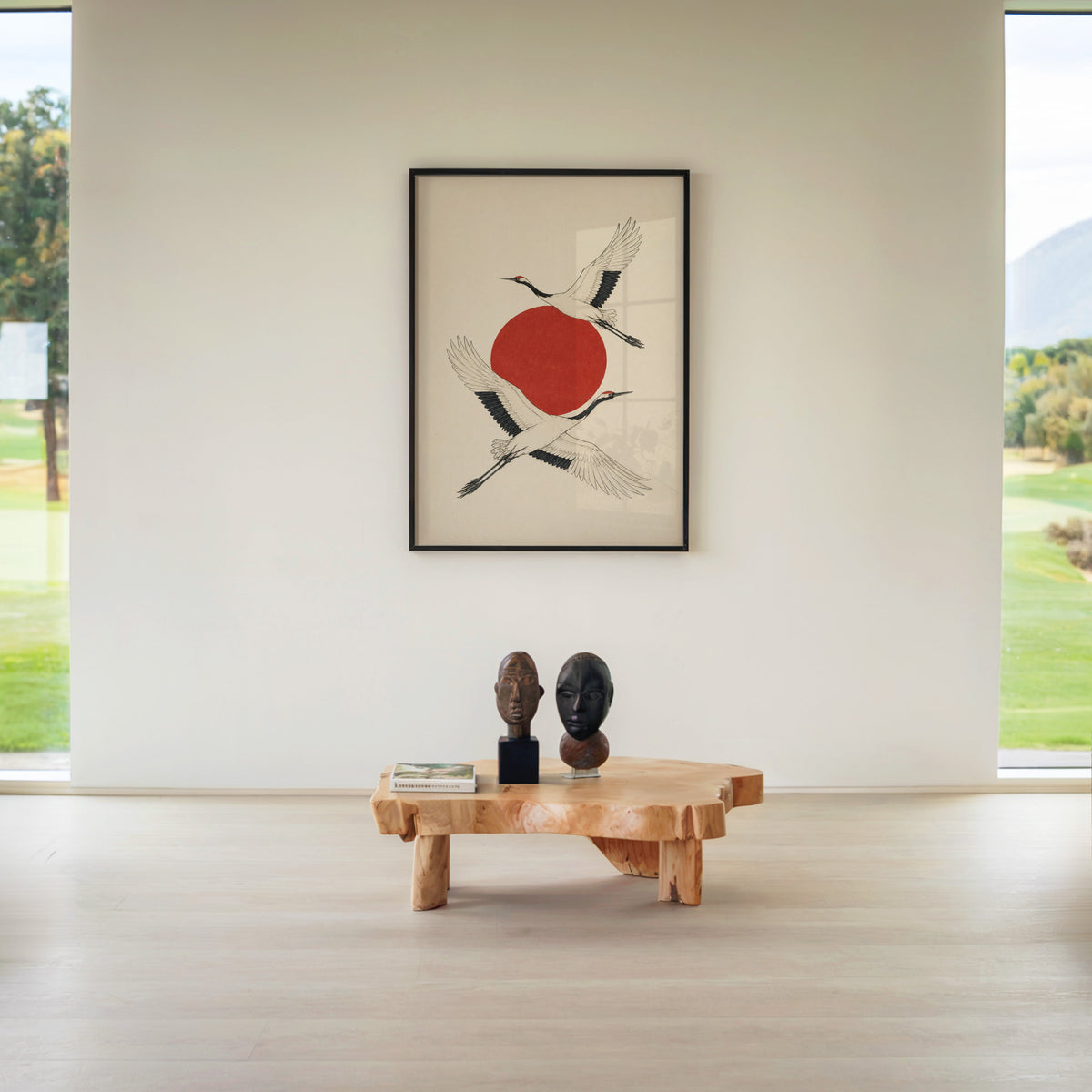 two cranes crossing a red sun print in a black frame above a live-edge wood coffee table between garden windows