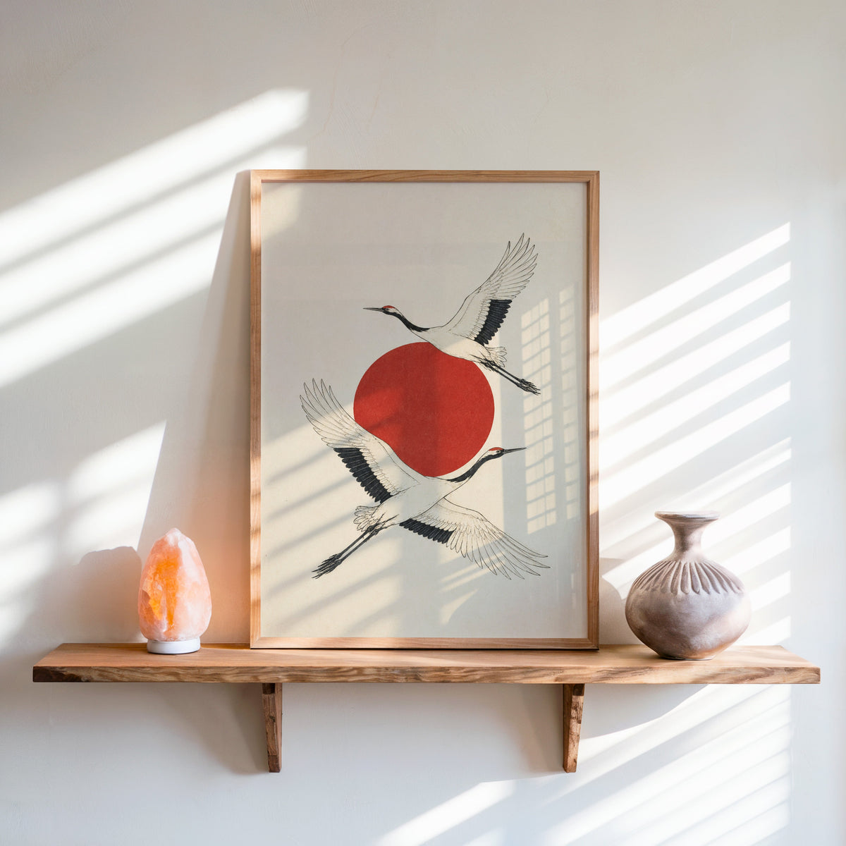 two cranes crossing a red sun print in an oak frame on a live-edge wall shelf with a salt lamp and clay vase