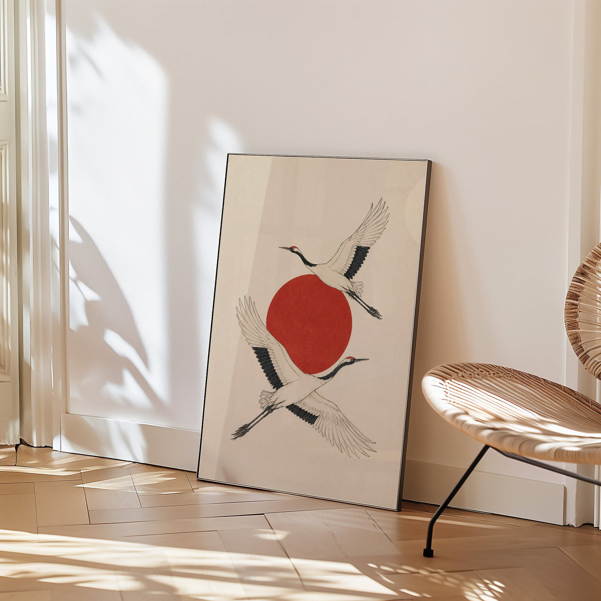 two cranes crossing a red sun print in a black frame leaning on a herringbone floor beside a woven rattan chair